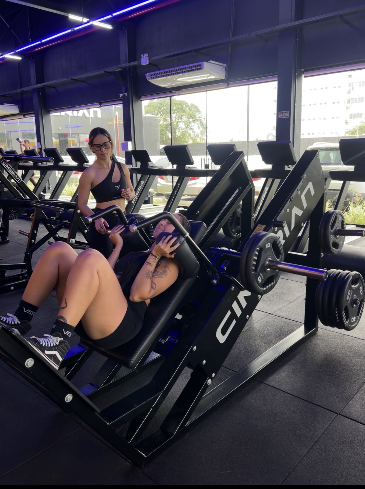 Aluna do Torres Squad treinando no hack squat com acompanhamento da equipe